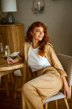 A person sitting on a table. The girl is sitting in the room at the table. She has long red hair. There is Christmas decor behind her. The girl is smiling, She has a wine glass in her hands