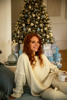 A woman sitting on a bed.A girl with red curly hair is sitting near the bed in the bedroom. Behind her in the room is a Christmas tree with toys and gifts. The girl is dressed in white knitted clothes