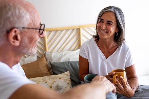 Happy mature woman drinking coffee in bed, having conversation with husband. Heterosexual couple concept.