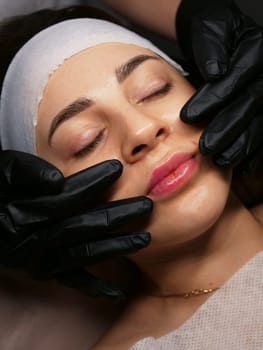 The beautician performs the procedure of microcurrents in black electric gloves to restore facial skin cells. Close-up, vertical