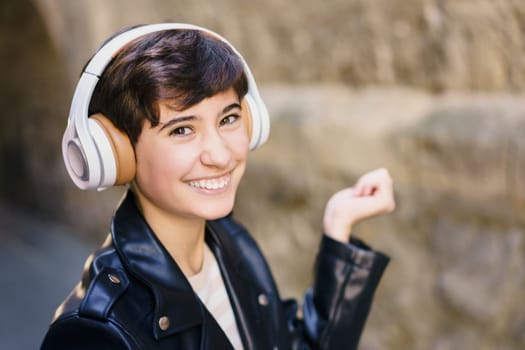 A cheerful young person is wearing trendy headphones while smiling broadly and making cheerful gestures in a vibrant urban setting, illustrating a deep passion for music and a carefree lifestyle