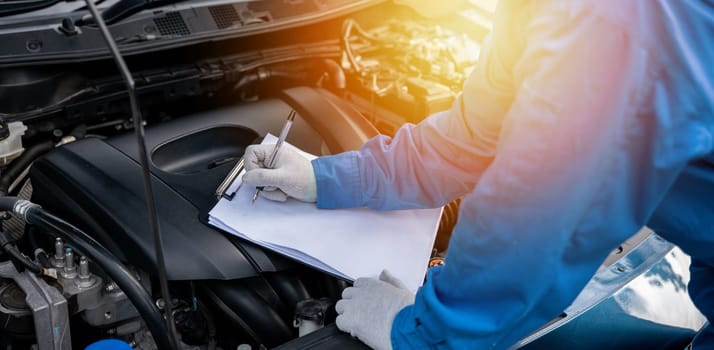 Skilled Asian mechanic in blue shirt writing on a paper while working on a car in an auto repair shop. Close-up shot of hand.