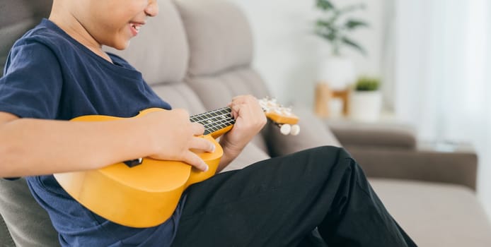 Little boy's hand strums the strings of a yellow ukulele on a couch. The cozy living room scene captures a moment of creativity and playful learning.