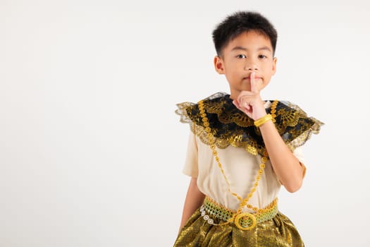 Portrait Thailand kid boy smiling traditional Thai dress costume making finger on lips mouth silent quiet gesture, studio isolated white background, primary child marking silence for stop quiet