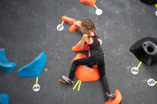 From below of determined girl in sportwear climbing artificial rock wall at bouldering gym. Low angle back view of sporty female child is training on equipment
