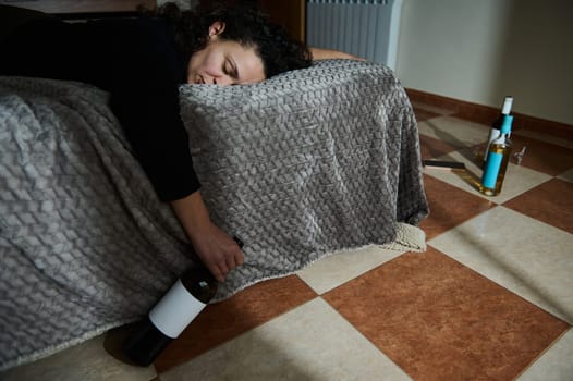 A person is lying on a sofa covered by a blanket holding a wine bottle with additional wine bottles placed on the floor.