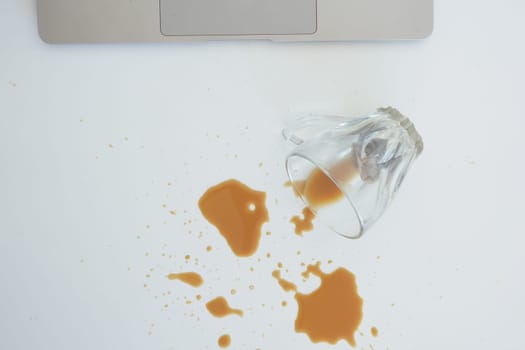 Overturned glass spilling drink on desk with laptop and glasses. Cleanup concept.