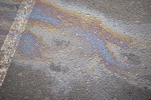 A vibrant oil spill spreads across the asphalt surface, reflecting a range of colors, contrasting with the white parking line. The scene captures an urban environment, highlighting pollution