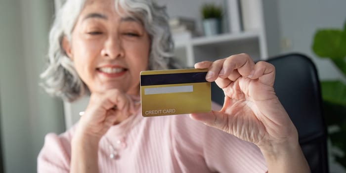 An elderly woman smiles brightly as she holds up her credit card, celebrating a successful online shopping experience in her comfortable home office.