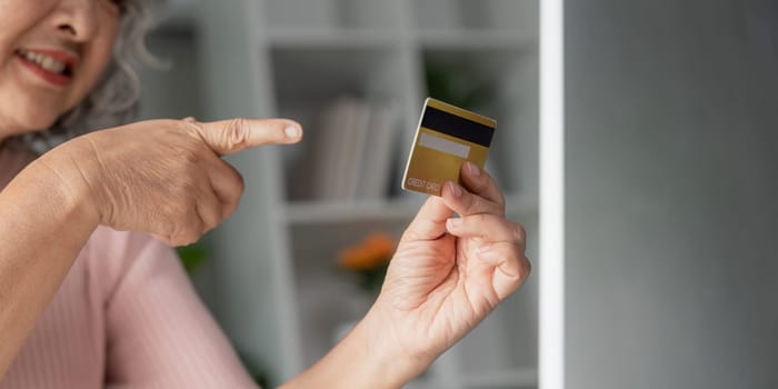 Enthusiastic elderly woman pointing at her credit card, highlighting her online shopping experience, within the comfort of her home office environment.
