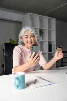 Joyful elderly woman using her smartphone to shop online, holding a credit card, sitting at her home office desk, exemplifying modern digital shopping.
