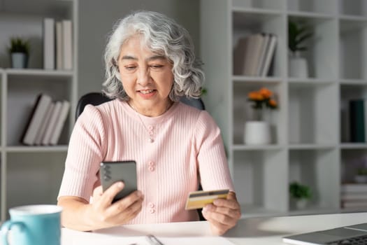 An elderly woman smiles while holding a credit card and using her smartphone for online shopping in a bright, inviting home office setting.