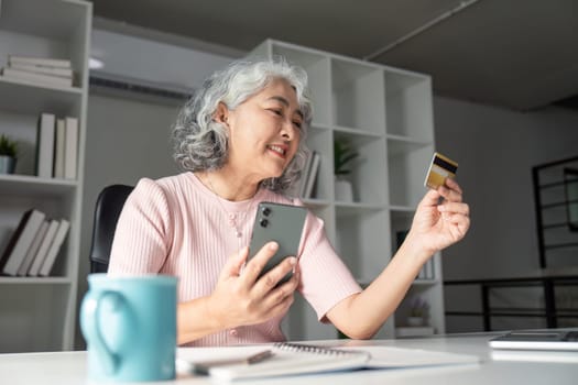 Smiling elderly woman shopping online, holding a credit card and smartphone while seated at her home office desk, showcasing the joy of digital shopping.