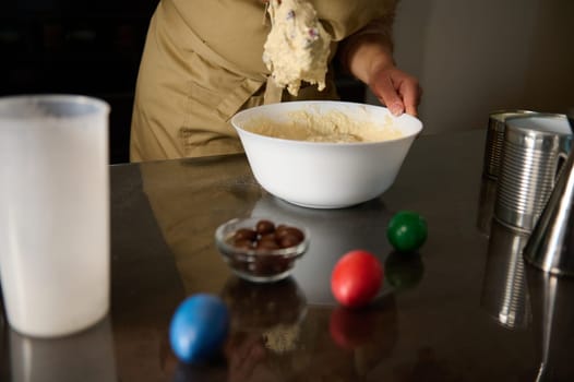 A person in an apron prepares dough in a bowl on a surface with colored eggs symbolizing Easter. The scene reflects holiday celebrations and homemade baking activities.