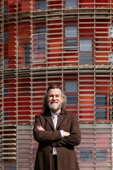 positive middle age male with beard in elegant suit standing with arms crossed and looking at camera against urban building, concept of business and mature man lifestyle, copyspace for text