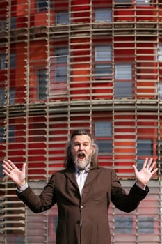 funny middle age male with beard in elegant suit surprised with arms and mouth open looking at camera against urban building, concept of business and mature man lifestyle, copyspace for text