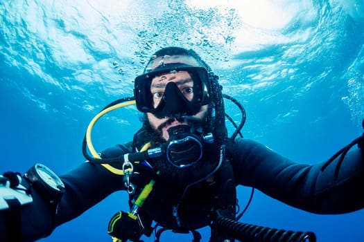 male scuba diver in black wetsuit and snorkeling mask looking at camera while taking selfie portrait in deep sea with clear water, concept of active leisure and underwater adventure