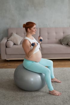 Pregnant woman doing exercises with dumbbells while sitting on a fitness ball at home