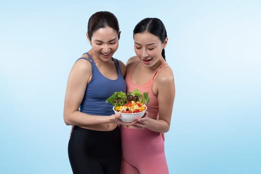 Two young sporty Asian women in sportswear holding salad bowl fill with fruit and vegetable. Natural youthful and fit body lifestyle people with balance nutrition on isolated background. Vigorous