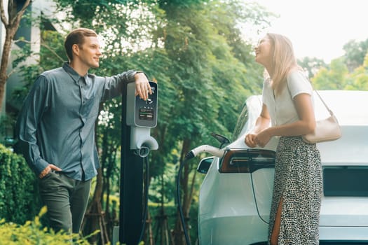 Young couple travel with EV electric car charging in green sustainable city outdoor garden in summer shows urban sustainability lifestyle by green clean rechargeable energy of electric vehicle innards