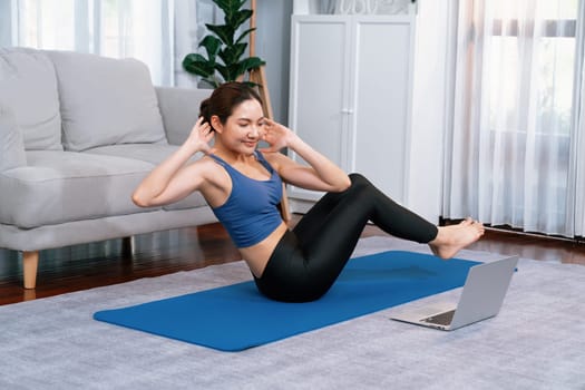Asian woman in sportswear doing crunch on exercising mat as home workout training routine. Attractive girl engage in her pursuit of healthy lifestyle with online exercise training video. Vigorous