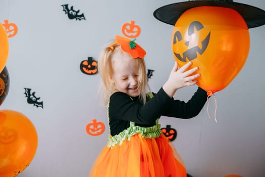 Children's Halloween - a girl in a witch hat and a carnival costume with airy orange and black balloons at home. Ready to celebrate Halloween.