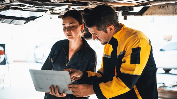 Two vehicle mechanic working together underneath lifted car, conduct car inspection with laptop. Automotive service technician in uniform carefully make diagnostic troubleshooting. Panorama Oxus