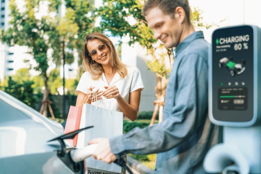 Young couple travel with EV electric car charging in green sustainable city outdoor garden in summer shows urban sustainability lifestyle by green clean rechargeable energy of electric vehicle innards