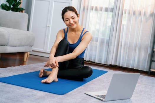 Asian woman in sportswear sitting on the house floor while watching online exercise training video on laptop. Attractive girl engage in her pursuit of healthy lifestyle. Vigorous