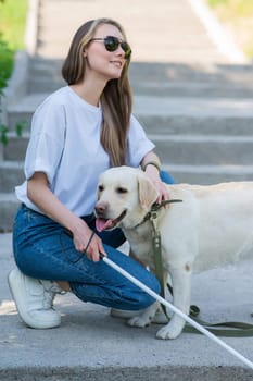 Blind young blonde petting a guide dog on a walk in the park. Woman with tactile cane at the stairs