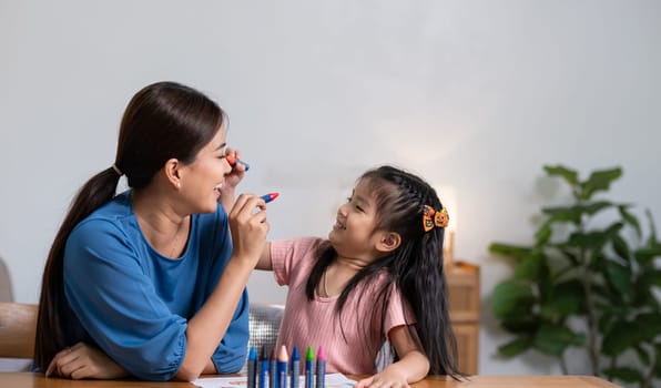 A young family spends their free time together in the living room at home. Mother and little daughter draw pictures with crayons on paper, smiling happy..