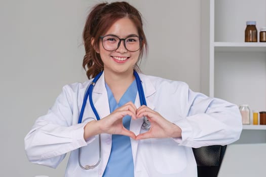 Beautiful asian woman wearing doctor uniform and stethoscope smiling in love doing heart symbol shape with hands. romantic concept..