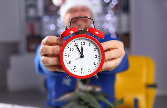 Bearded smiling man wearing warm blue sweater with traditional deer hold arm red alarm clock. Within inch coming 2024 year with glowing garland background concept closeup