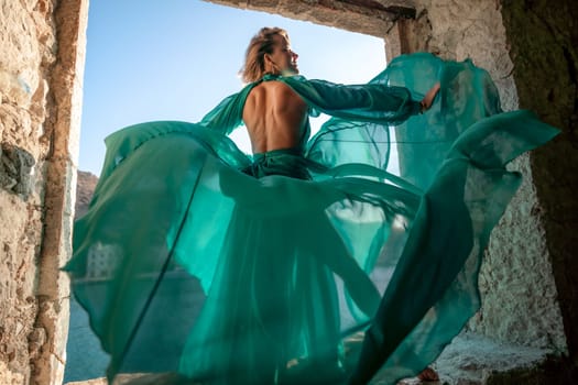 Rear view of a happy blonde woman in a long mint dress posing against the backdrop of the sea in an old building with columns. Girl in nature against the blue sky