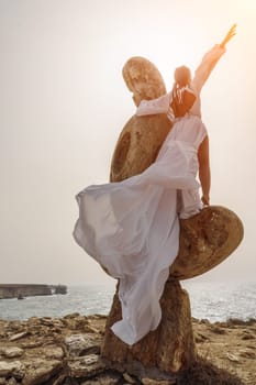 Woman sky stone. A woman stands on a stone sculpture made of large stones. She is dressed in a white long dress, against the backdrop of the sea and sky. The dress develops in the wind
