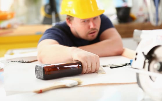 Arm of drunken worker in yellow helmet hold liquor bottle sleeping at table closeup. Manual job workplace DIY inspiration fix shop hard hat industrial education profession career