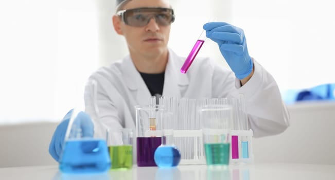 A male chemist holds test tube of glass in his hand overflows a liquid solution of potassium permanganate. Conducts an analysis reaction takes various versions of reagents using chemical manufacturing.