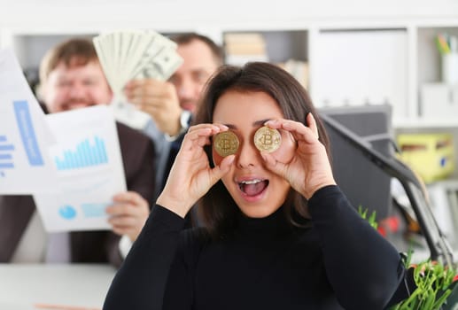 Young happy smiling chinese woman holding two coins of bitcoins in hand instead of glasses rejoices that she had time to buy to produce. Moment deshego before the rise in price and growth.