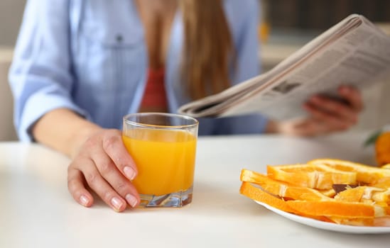 Young beautiful attractive girl in the kitchen reading a newspaper with news during breakfast and drinking orange juice holding a glass in her hand