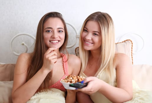 Portrait of two beautiful young smiling woman friends watching a comedy movie melodramma. In an apartment eating popcorn and laughing on the bed in pajamas during a hen party switch channels tv.