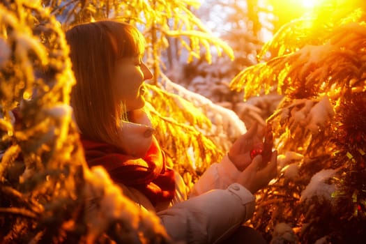 A middle-aged woman strolls through a snowy forest, admiring the beauty of winter while holding a festive ornament in her hands.