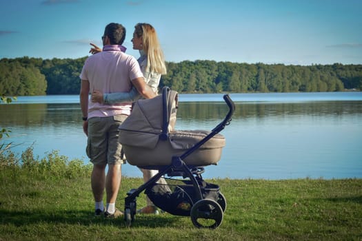 Young couple walking with newborn baby in stroller on lake summer day.