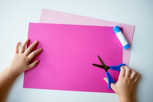 Childs hands crafting with pink paper, blue scissors, and glue on light background.