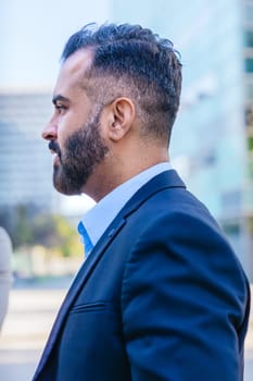 Side profile of a professionally dressed businessman with a beard, standing outdoors in an urban environment with modern buildings in the background.