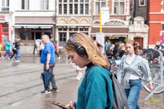 young student girl in headphones and blue sweater uses smartphone while waiting for bus in city among other people,unlimited internet access, street after rain, Ghent, Belgium, September 24, 2024,