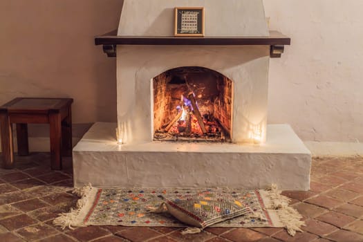 A warm and inviting scene featuring a lit fireplace with glowing firewood and candles. A colorful traditional rug and pillow lie on the floor, creating a cozy atmosphere. Home, relaxation, and rustic comfort concept.
