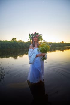 Slavic plump chubby girl in long white dress on the feast of Ivan Kupala with flowers and water in a river or lake on summer evening