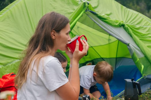 A woman willingly enjoys a refreshing drink while her children happily play in a vibrant tent during a camping trip