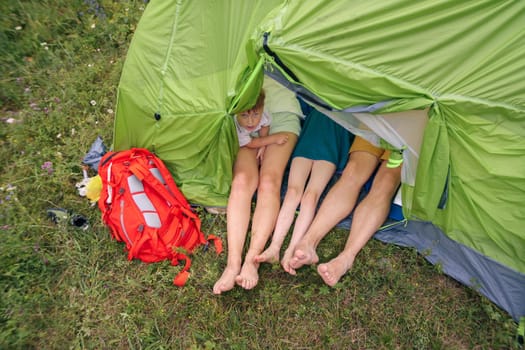 Three pairs of feet are protruding from a green tent, with backpacks resting nearby on a bright, sunny camping day