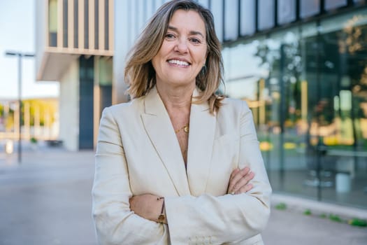 Portrait of a young businesswoman in a beige blazer standing outdoors in an urban setting with arms crossed, exuding confidence and professionalism
